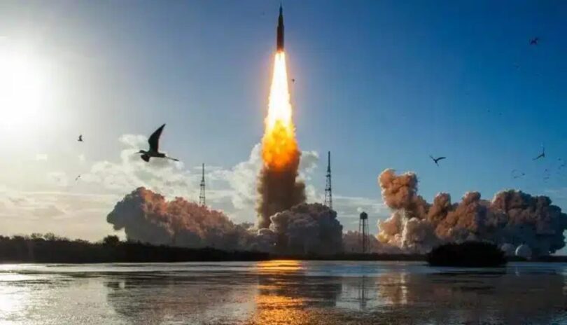 Photographie du décollage de la fusée SLS de la NASA depuis le Kennedy Space Center pour la mission Artemis II, montrant une colonne de feu intense et de la fumée s'élevant au-dessus d'un plan d'eau avec des oiseaux en vol au premier plan.