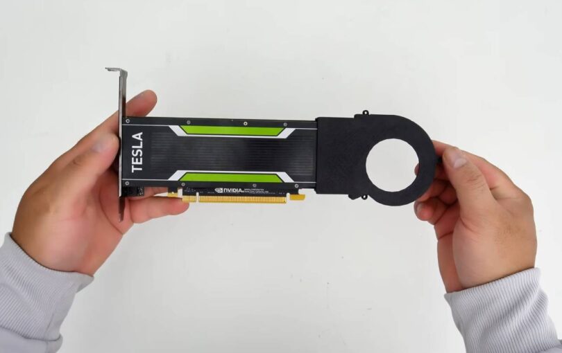 Vue de dessus d'une carte graphique Nvidia Tesla low-profile, modifiée avec un carénage de ventilateur passif noir imprimé en 3D, tenue en main par une personne portant un pull gris sur un fond blanc.