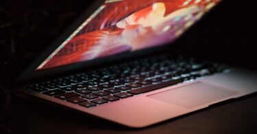 Vue latérale d'un ordinateur portable MacBook ouvert avec un écran rétroéclairé coloré dans un environnement sombre, illustrant un utilisateur en attente de mise à jour système.