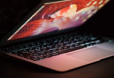 Vue latérale d'un ordinateur portable MacBook ouvert avec un écran rétroéclairé coloré dans un environnement sombre, illustrant un utilisateur en attente de mise à jour système.