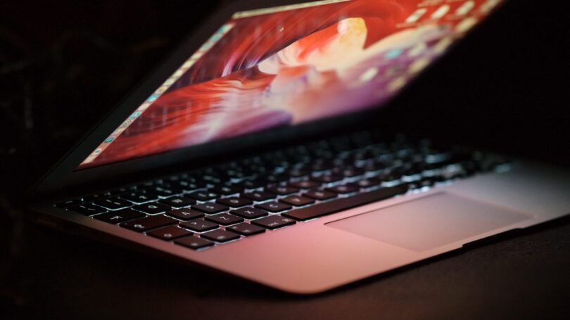 Vue latérale d'un ordinateur portable MacBook ouvert avec un écran rétroéclairé coloré dans un environnement sombre, illustrant un utilisateur en attente de mise à jour système.