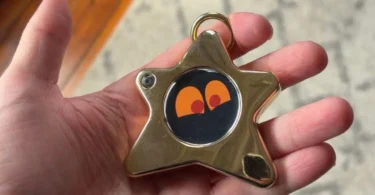Gros plan sur le Starboy, un porte-clés connecté en forme d'étoile dorée avec un écran OLED affichant des yeux expressifs orange, tenu dans une main.