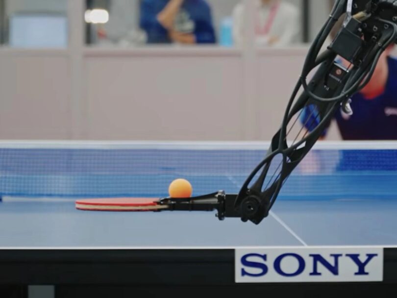 Gros plan d'un bras robotique Sony équipé d'une raquette de tennis de table, s'apprêtant à frapper une balle orange avec une précision millimétrée sur une table officielle.