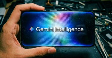 Smartphone tenu en main affichant le logo Gemini Intelligence de Google sur un fond étoilé et lumineux, posé sur un plan de travail électronique en vue de l'intégration dans Android 17.