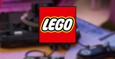 Logo officiel LEGO superposé en premier plan sur un arrière-plan flou montrant une console de jeux rétro noire avec ses manettes filaires sur une table en bois.