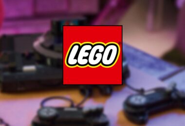 Logo officiel LEGO superposé en premier plan sur un arrière-plan flou montrant une console de jeux rétro noire avec ses manettes filaires sur une table en bois.