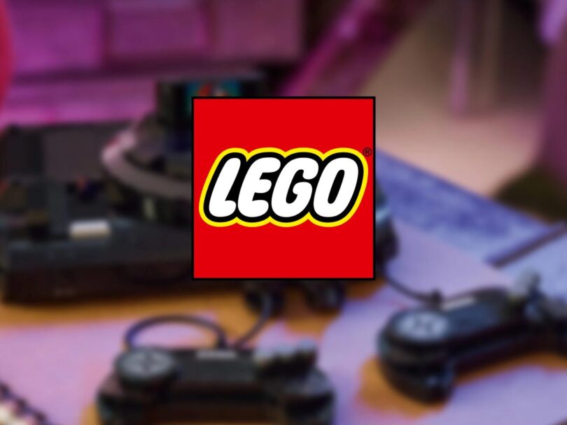 Logo officiel LEGO superposé en premier plan sur un arrière-plan flou montrant une console de jeux rétro noire avec ses manettes filaires sur une table en bois.