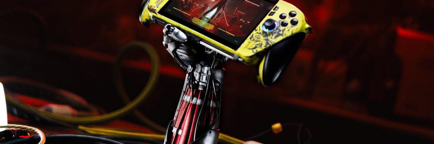 Console Asus ROG Xbox Ally X personnalisée aux couleurs de Cyberpunk 2077 (jaune vieilli), maintenue par un support exclusif en forme de bras cybernétique de Johnny Silverhand, entourée de câbles.