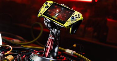 Console Asus ROG Xbox Ally X personnalisée aux couleurs de Cyberpunk 2077 (jaune vieilli), maintenue par un support exclusif en forme de bras cybernétique de Johnny Silverhand, entourée de câbles.