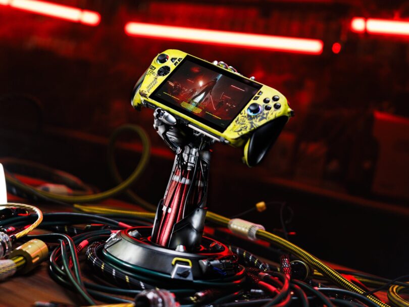 Console Asus ROG Xbox Ally X personnalisée aux couleurs de Cyberpunk 2077 (jaune vieilli), maintenue par un support exclusif en forme de bras cybernétique de Johnny Silverhand, entourée de câbles.