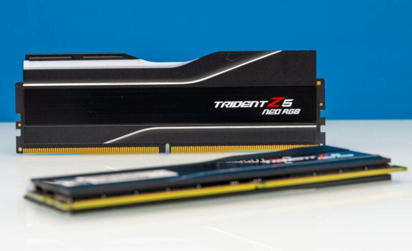 Deux barrettes de mémoire vive G.Skill Trident Z5 RGB Neo DDR5 noires avec dissipateur thermique en aluminium brossé et diffuseur LED RGB, posées sur une surface blanche devant un fond bleu.